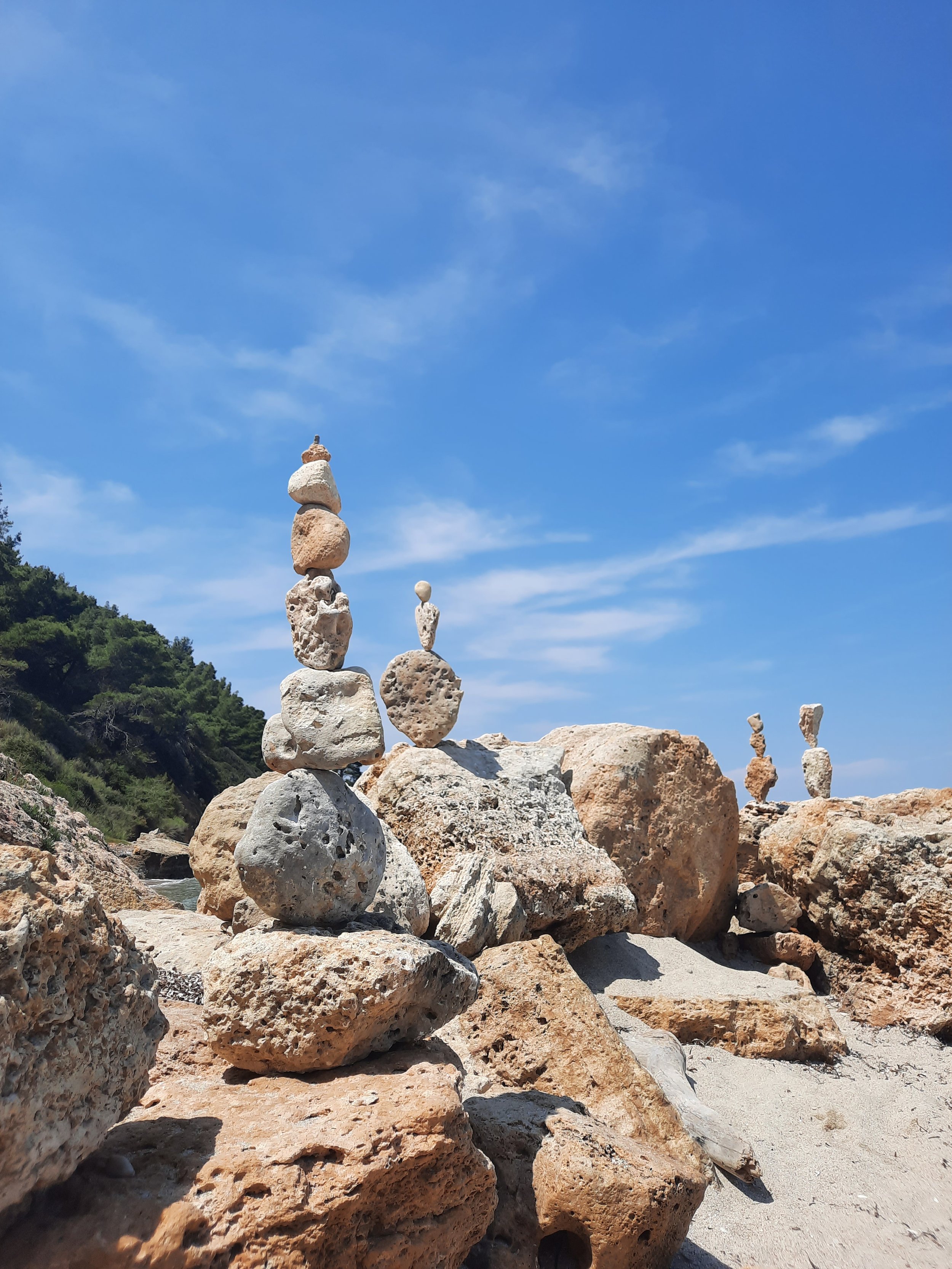 Steinadationen am Strand unter blauem Himmel, mit Kies und Sand, im Hintergrund grüne Hügel.