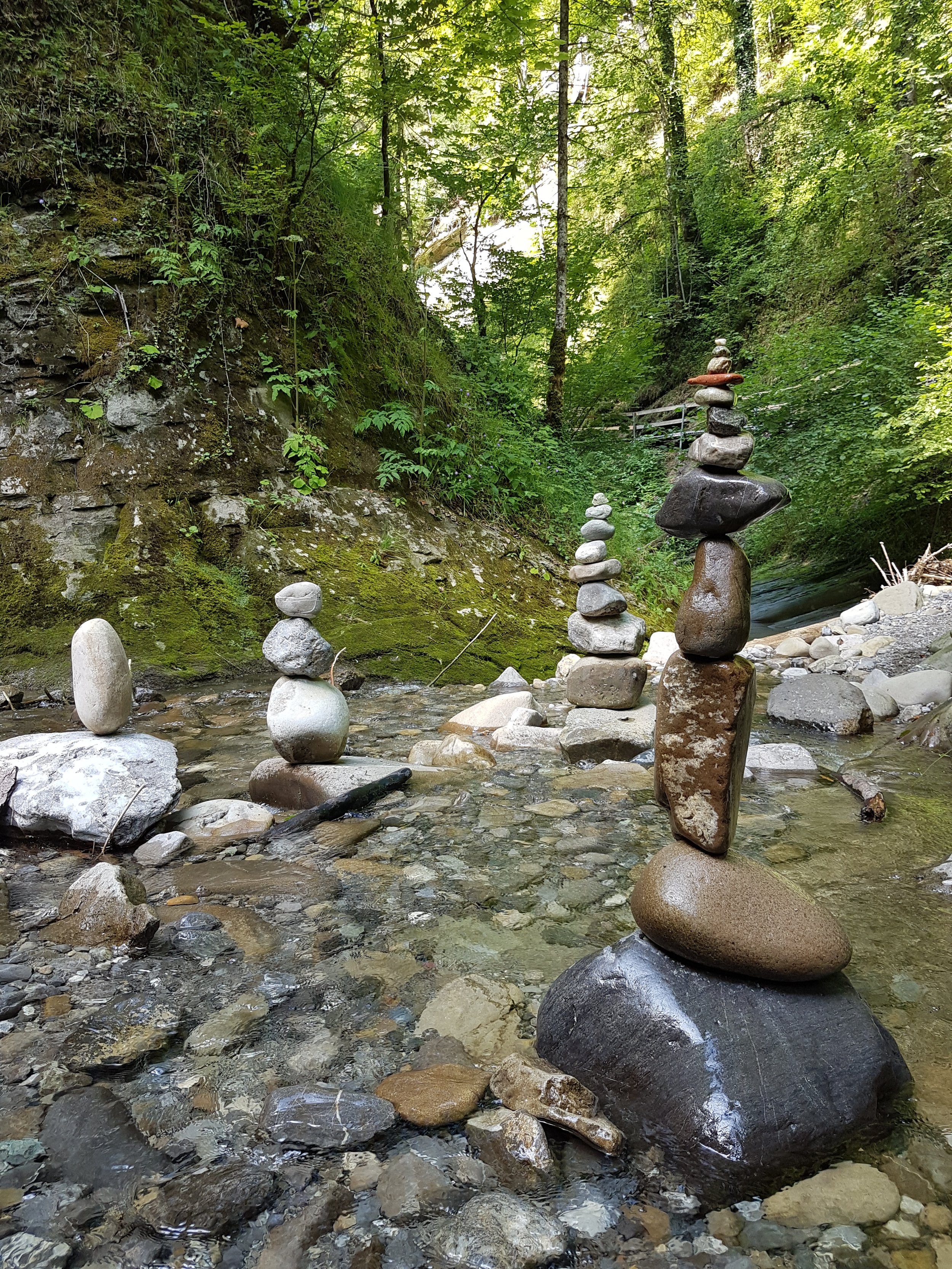 Steinshaufen in einem Fluss in einem grünen Wald, mit einem Wanderweg und einem Holzsteg im Hintergrund.