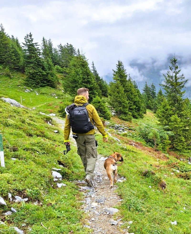 Ein Mann mit Rucksack und Hund wandert auf einem schmalen Pfad durch eine grüne, bergige Landschaft mit Nadelbäumen und Wolken am Himmel.