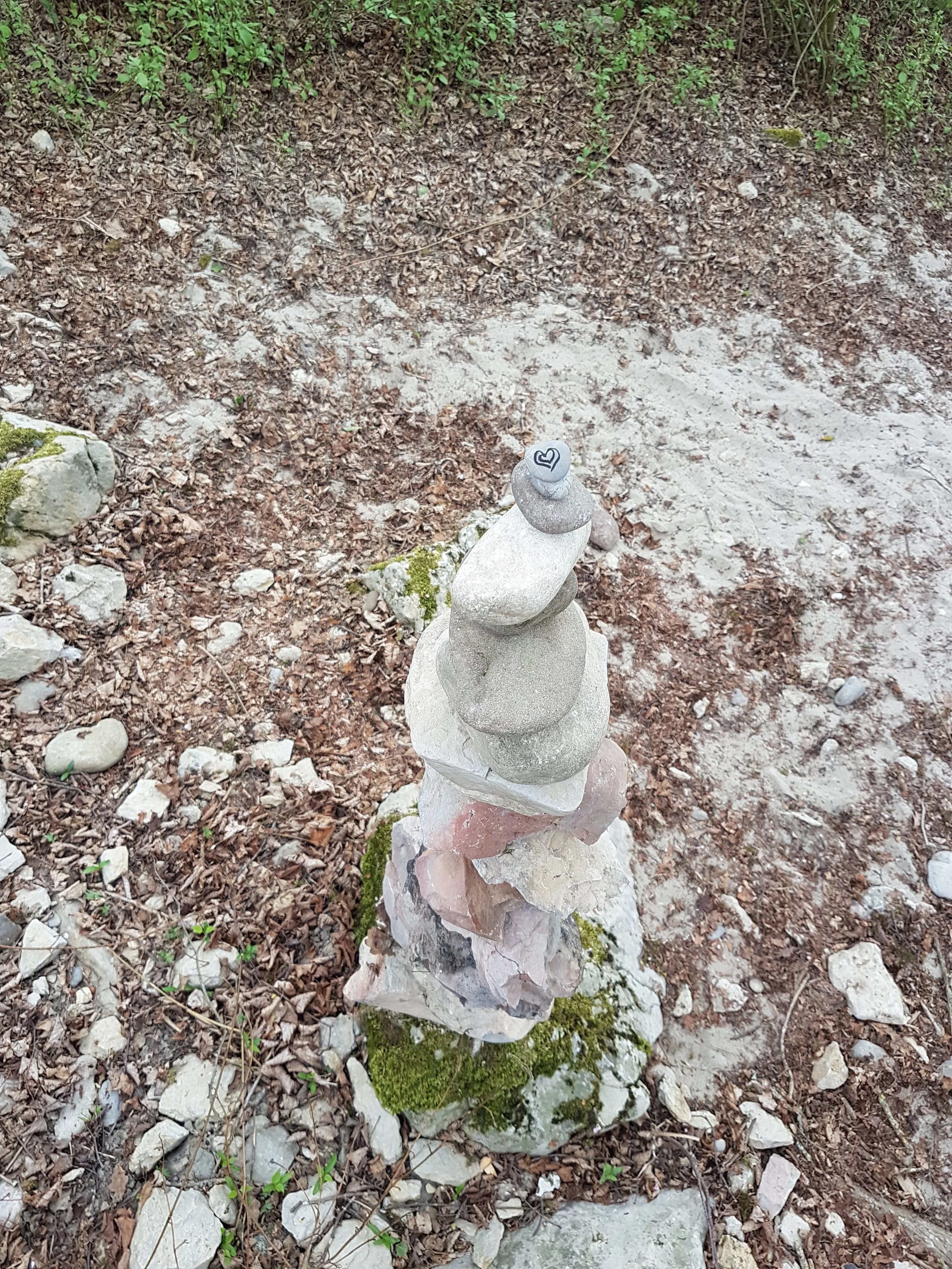 Ein steinerner Turm aus verschiedenen Steinen auf glattem Untergrund, umgeben von trockenem Boden und kleineren Steinen, mit grünem Moos am Sockel.