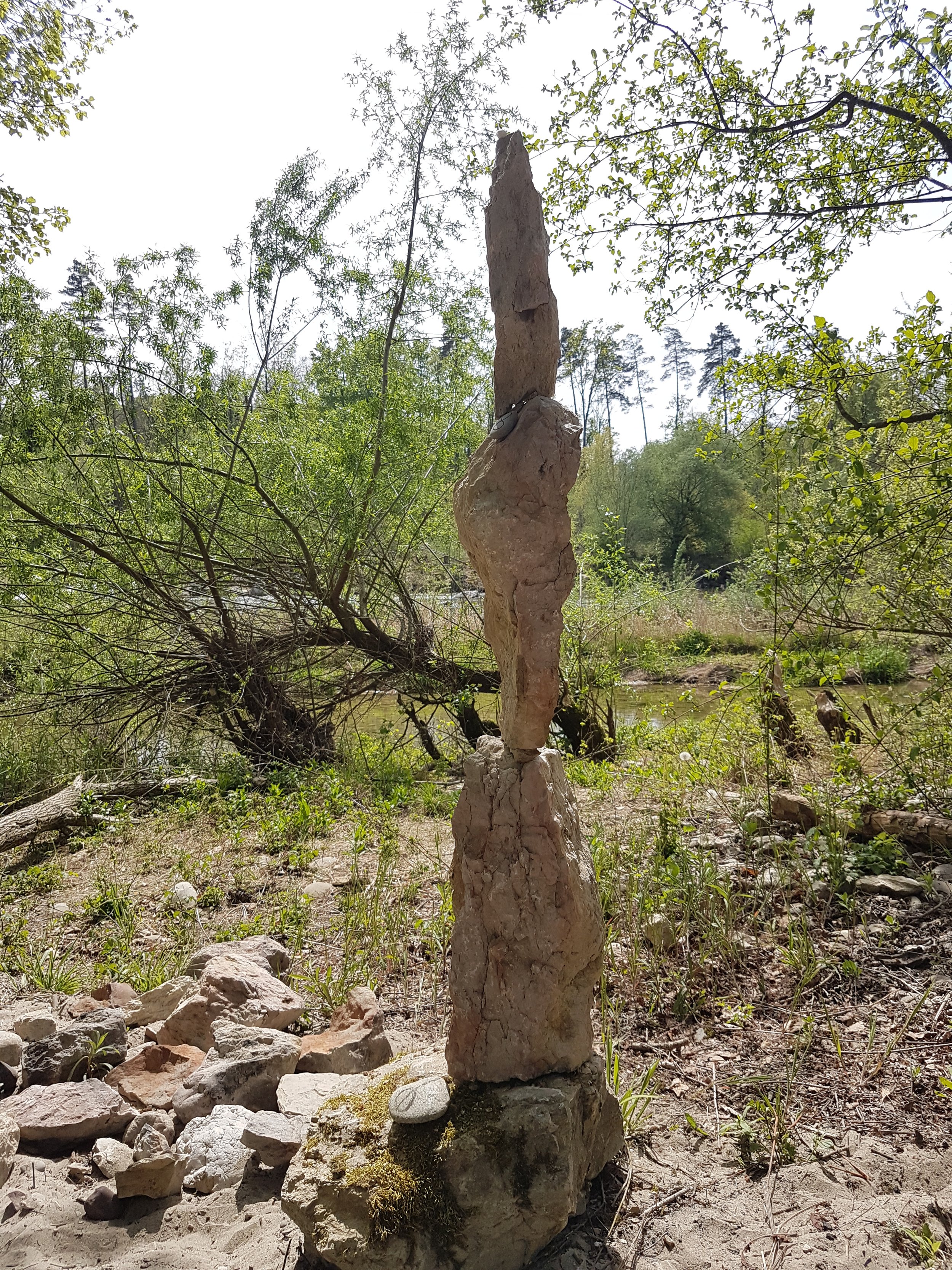 Kunstvoller Steinturm in einer natürlichen Umgebung mit Bäumen und Wasser im Hintergrund.