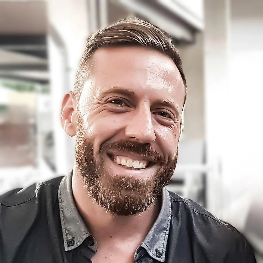 Ein Mann mit braunem Bart und kurzen braunen Haaren lächelt in die Kamera, im Hintergrund eine Küche.