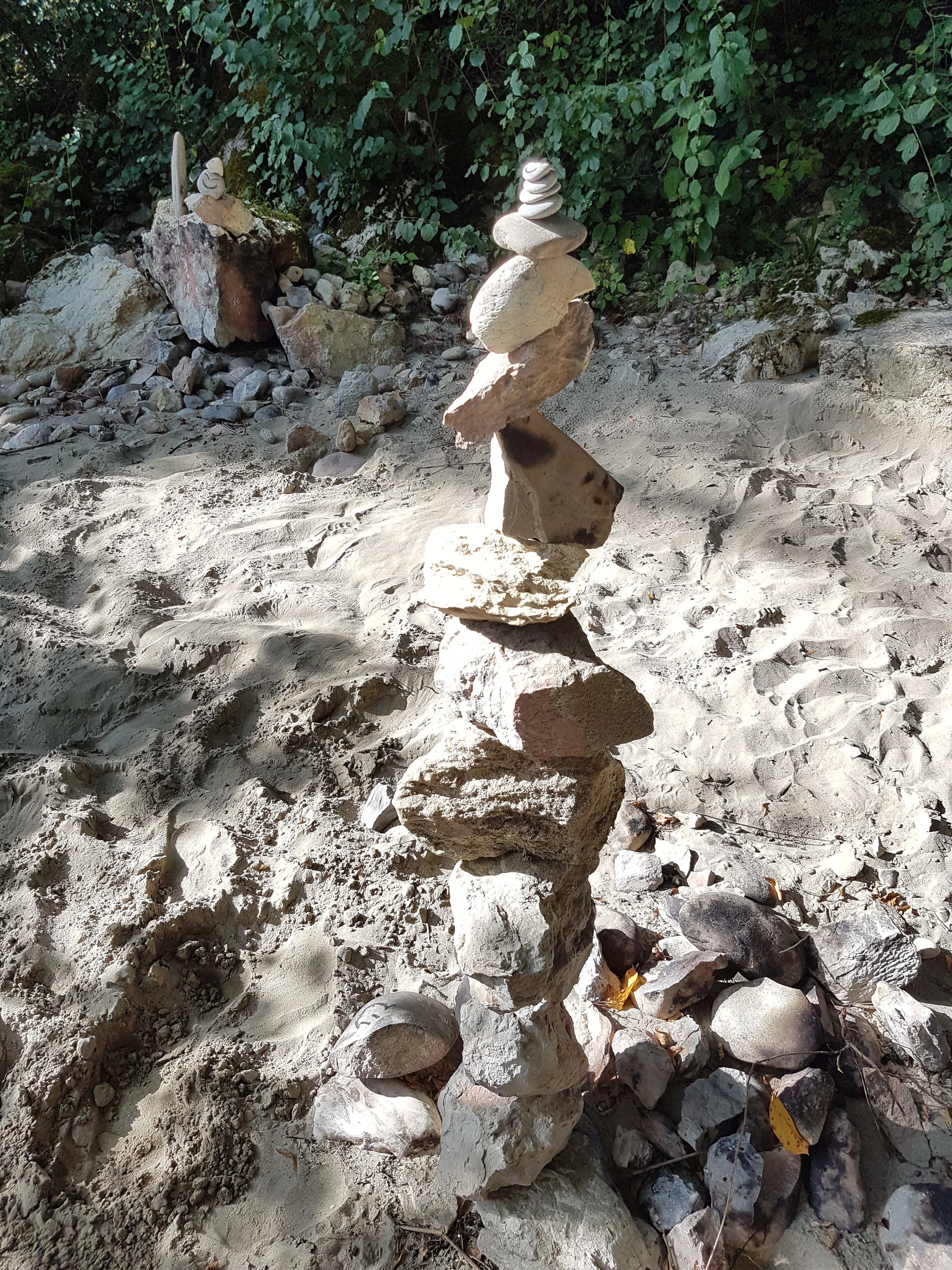 Sandsteinstapel mit mehreren gestapelten Steinen verschiedener Größen und Formen, auf sandigem Boden mit einigen Blättern, im Hintergrund dichter grüner Busch.
