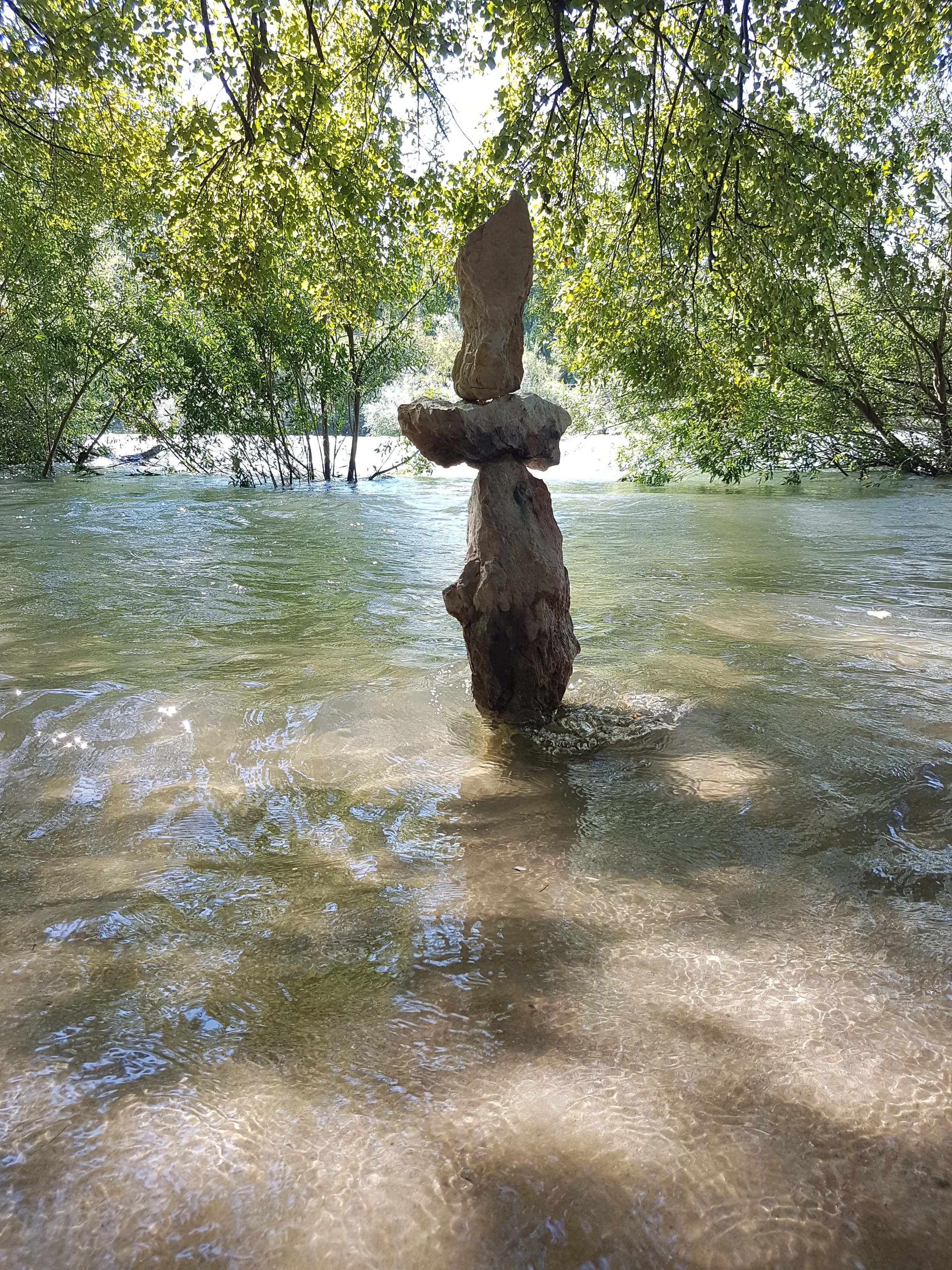 Ein Wasser füllter Fluss oder See mit einem kleinen Steinmännchen in der Mitte, bestehend aus mehreren Steinen, umgeben von grünen Bäumen und Blättern.