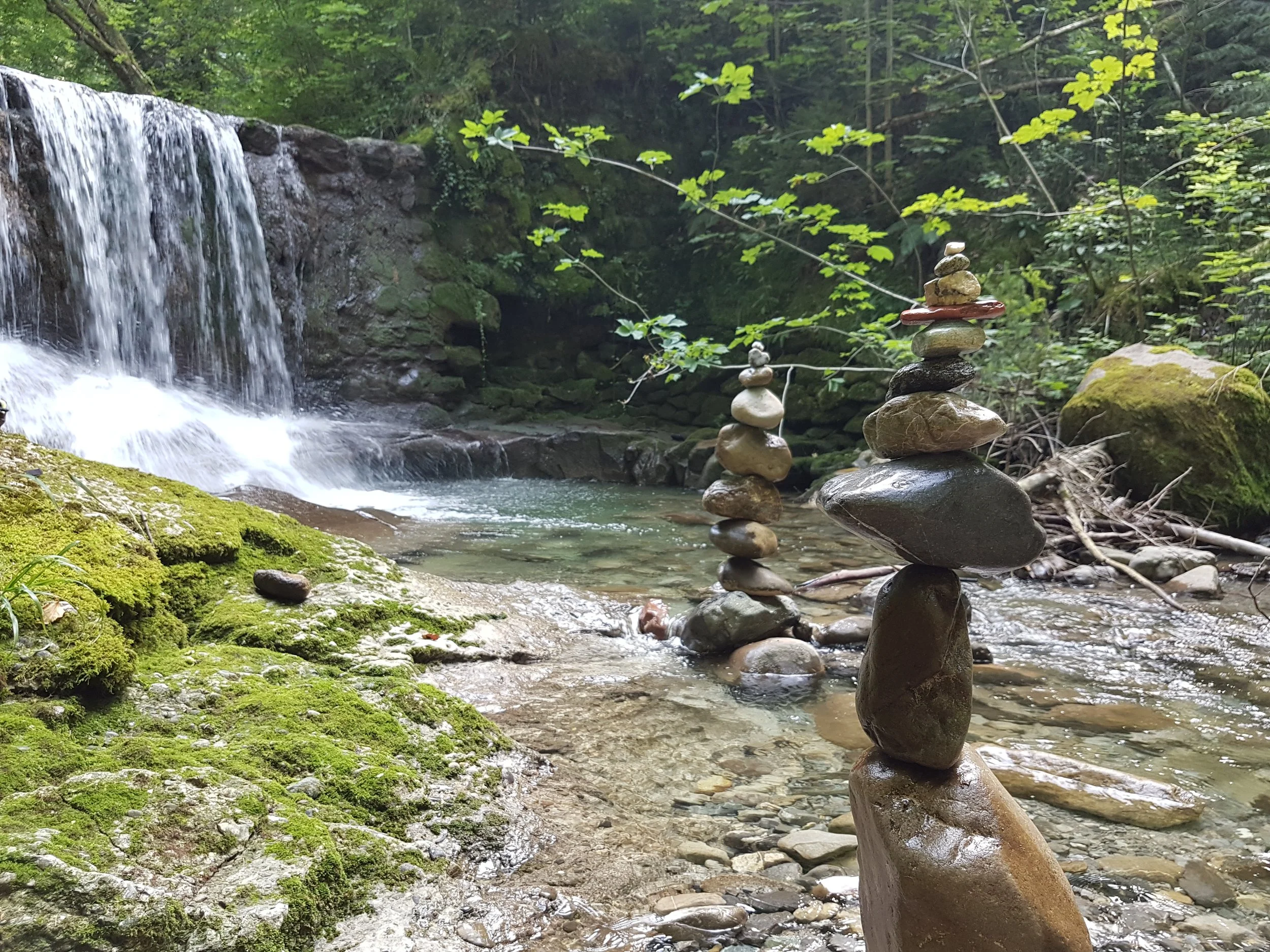 Felsen im Fluss, einige kunstvoll aus Steinen aufgestapelt, im Hintergrund ein kleiner Wasserfall in einem grünen Wald.