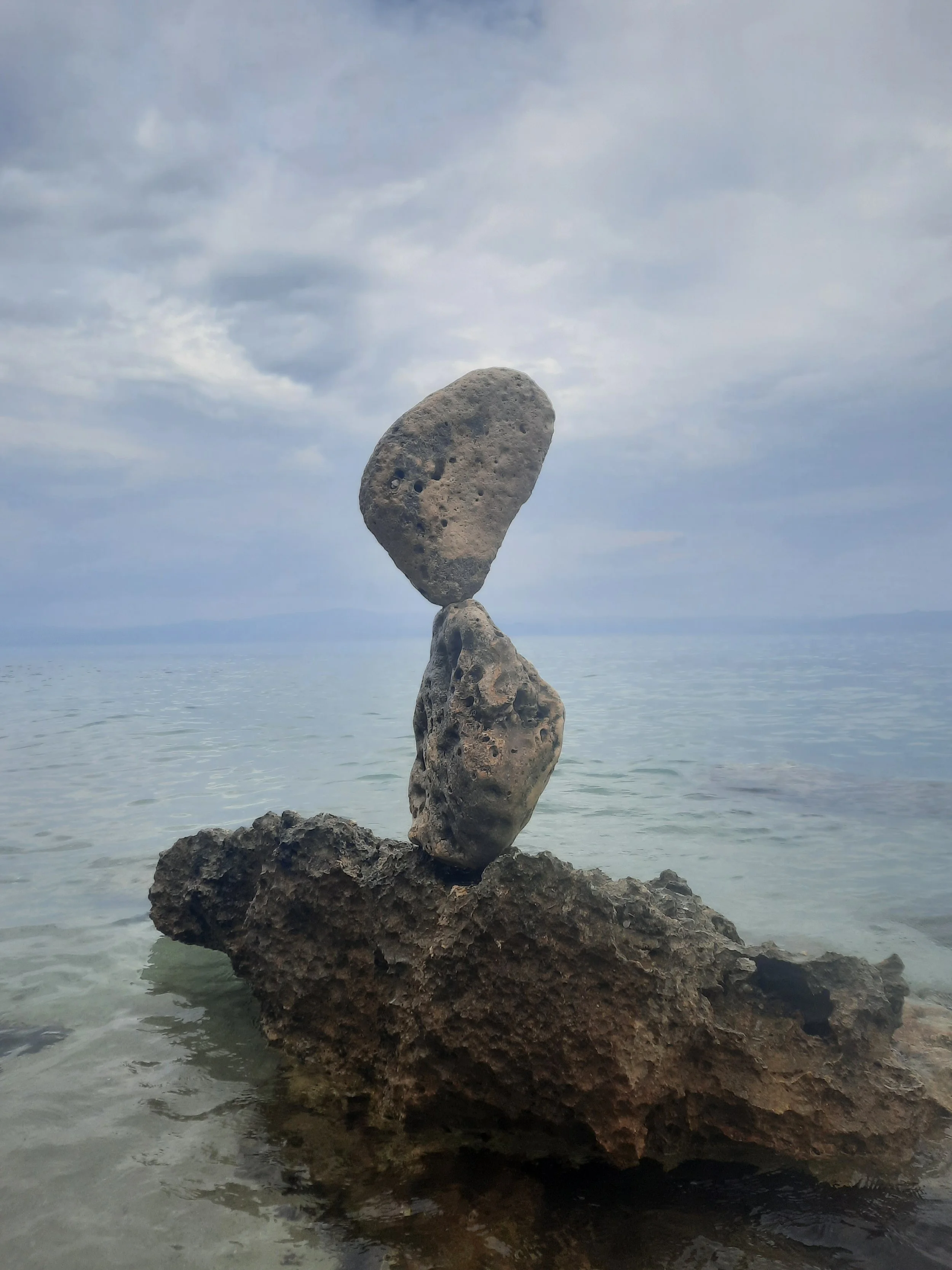 Zwei Steine kunstvoll auf einem größeren Felsen im Wasser balanciert, vor bewölktem Himmel mit Meer im Hintergrund.