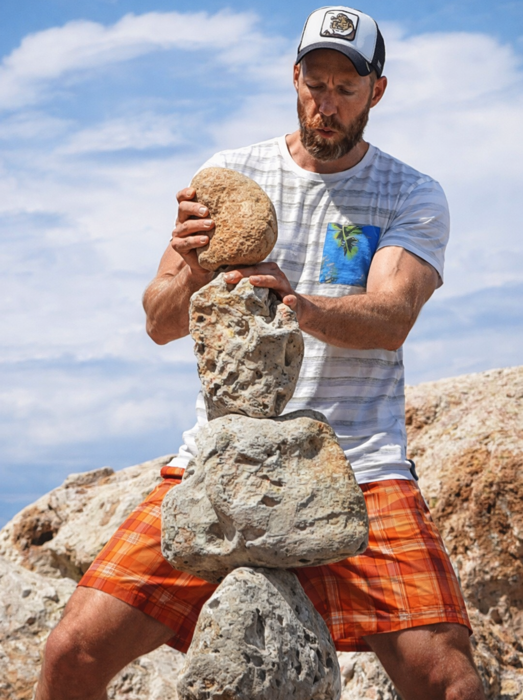 Mann in sommerlicher Kleidung baut einen Steinturm an einem Strand auf, hinter ihm Himmel mit wenigen Wolken.