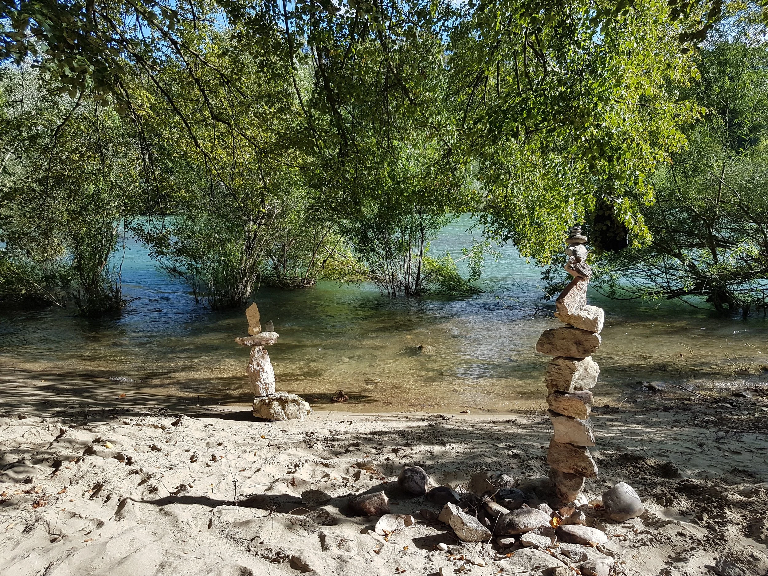 Zwei Steinkunstwerke am Ufer eines Flusses, umgeben von Bäumen und Wasser.