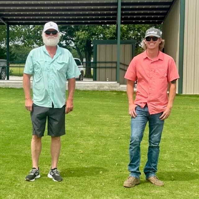 Two men standing outdoors on a grassy area, smiling and wearing casual summer clothes and sunglasses. One man has a white beard, wearing a light green short-sleeve shirt, dark shorts, sneakers, and a white cap. The other man has curly hair, wearing a pink short-sleeve shirt, blue jeans, boots, and a cap.