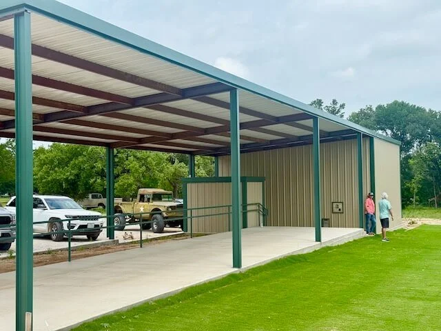 A portable shelter with a metal roof and open sides next to a grassy area, with two women standing nearby and cars parked in the background.