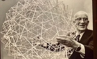 A smiling man in a suit holding a complex geometric structure made of interconnected sticks or straws.