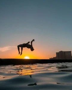 Person mid-air diving into water at sunset with building silhouette in the background.