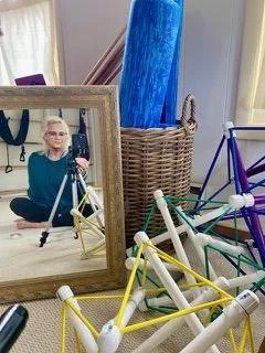 Reflection of Elizabeth Dare Andes taking a mirror selfie, surrounded by colorful geometric models and artistic objects.
