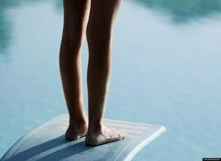 Person standing on a diving board on calm water, view of legs and feet.