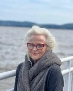A woman with white hair and glasses standing outdoors near a body of water, wearing a dark coat and a gray scarf.