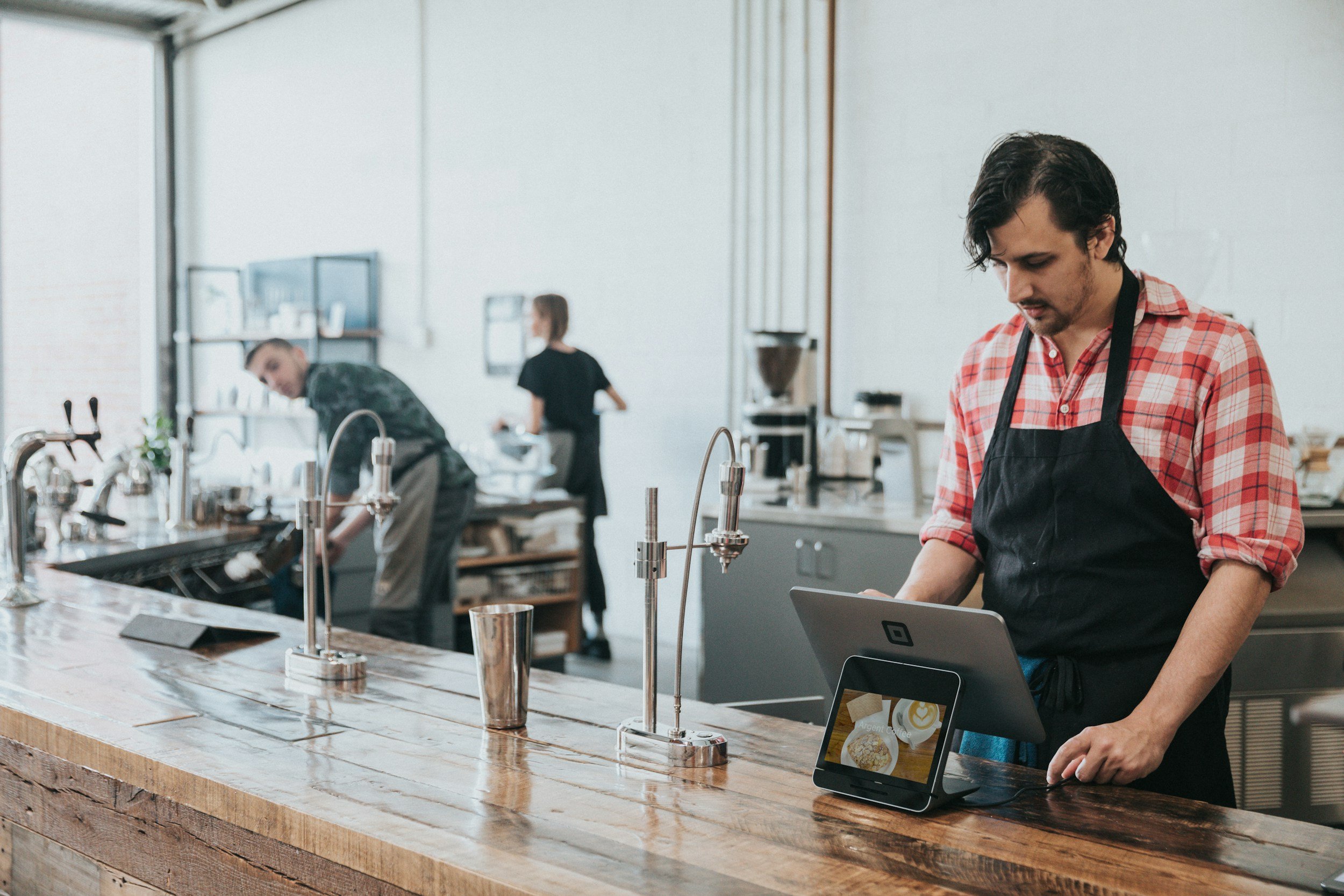 A man in a red plaid shirt and black apron standing behind a wooden counter with a tablet and a small monitor in a modern coffee shop or cafe. In the background, two other employees are working, one washing dishes and another preparing items.