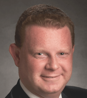 Close-up of a smiling man with curly brown hair and fair skin, wearing a dark suit, against a gray background.