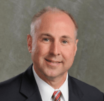 Portrait of a middle-aged man with short gray hair, wearing a black suit, white shirt, and striped tie, smiling against a grey background.