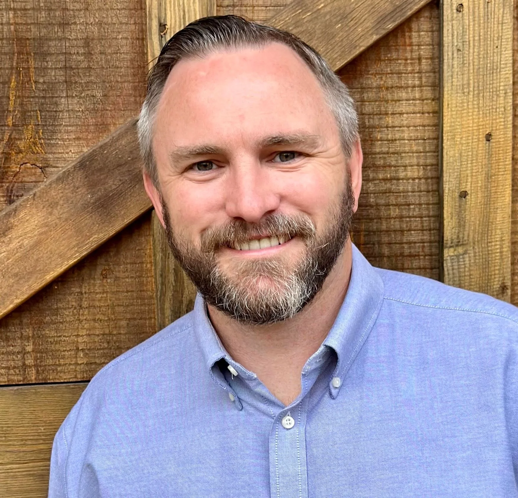 A man with a beard in a light blue button-up shirt smiling in front of a wooden fence.