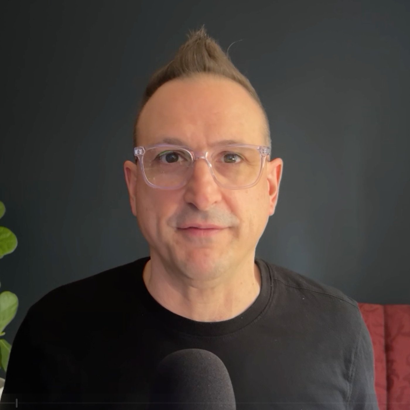 A man with clear glasses and a unique hairstyle facing the camera with a dark background.