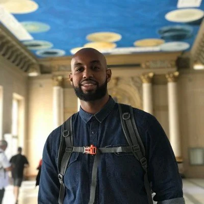 Man with backpack standing indoors with ornate columns and a painted ceiling in the background.