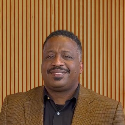 A man with short curly hair and a mustache wearing a black shirt and brown blazer, smiling at the camera, standing in front of a wooden slatted wall.