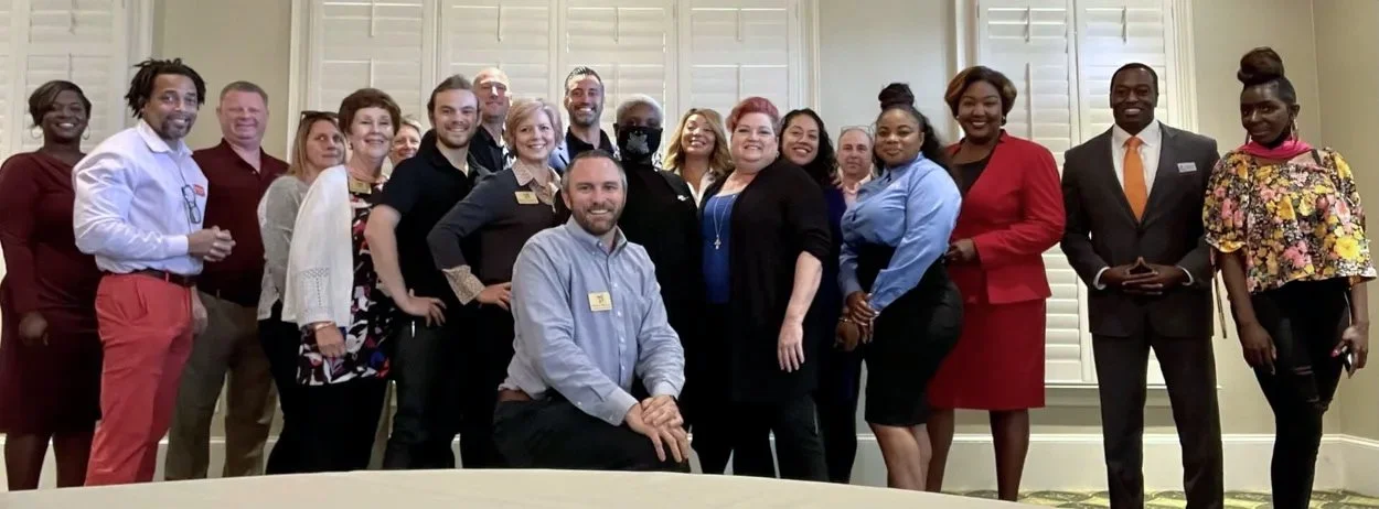 A diverse group of professionally dressed people posing together indoors, smiling for the camera.