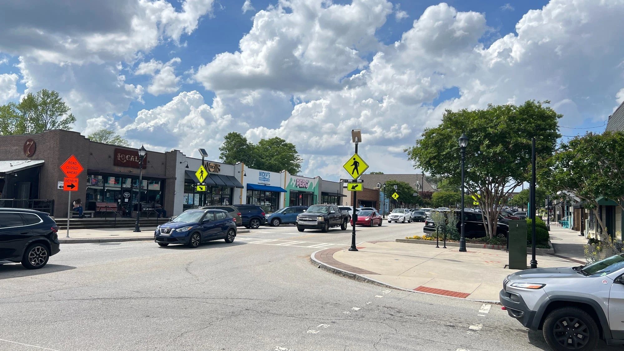 A small shopping district with parked cars, storefronts, pedestrian crosswalk signs, and a cloudy sky.