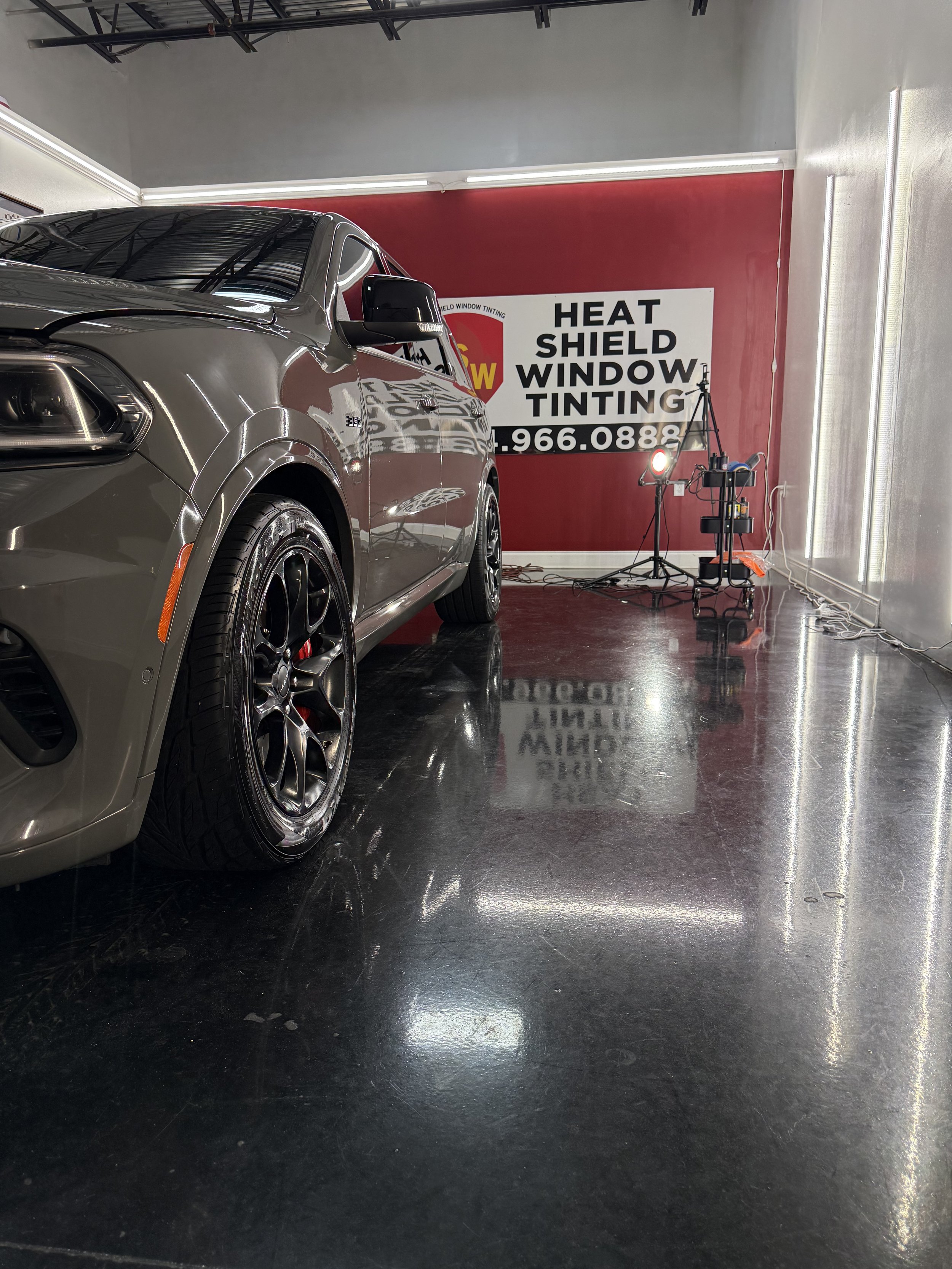 A tan car parked inside a garage or showroom with a red wall in the background. The wall has a sign reading 'HEAT SHIELD WINDOW TINTING' with a phone number. There is lighting equipment and cords on the floor near the wall.