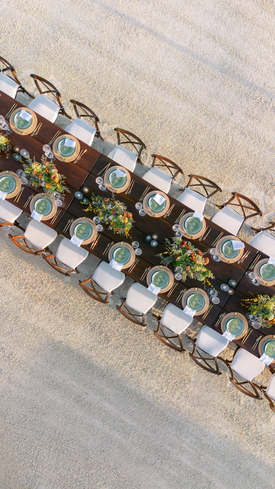 Overhead view of a long dining table set for a formal outdoor event on the beach, with white chairs, floral centerpieces, and place settings.