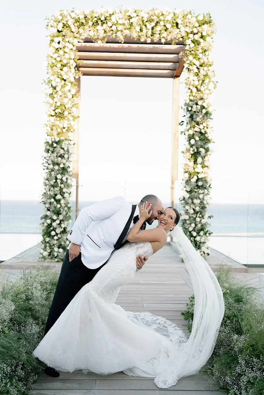 A bride and groom dancing together at an outdoor wedding ceremony on a deck overlooking the ocean, with a floral arch above them.