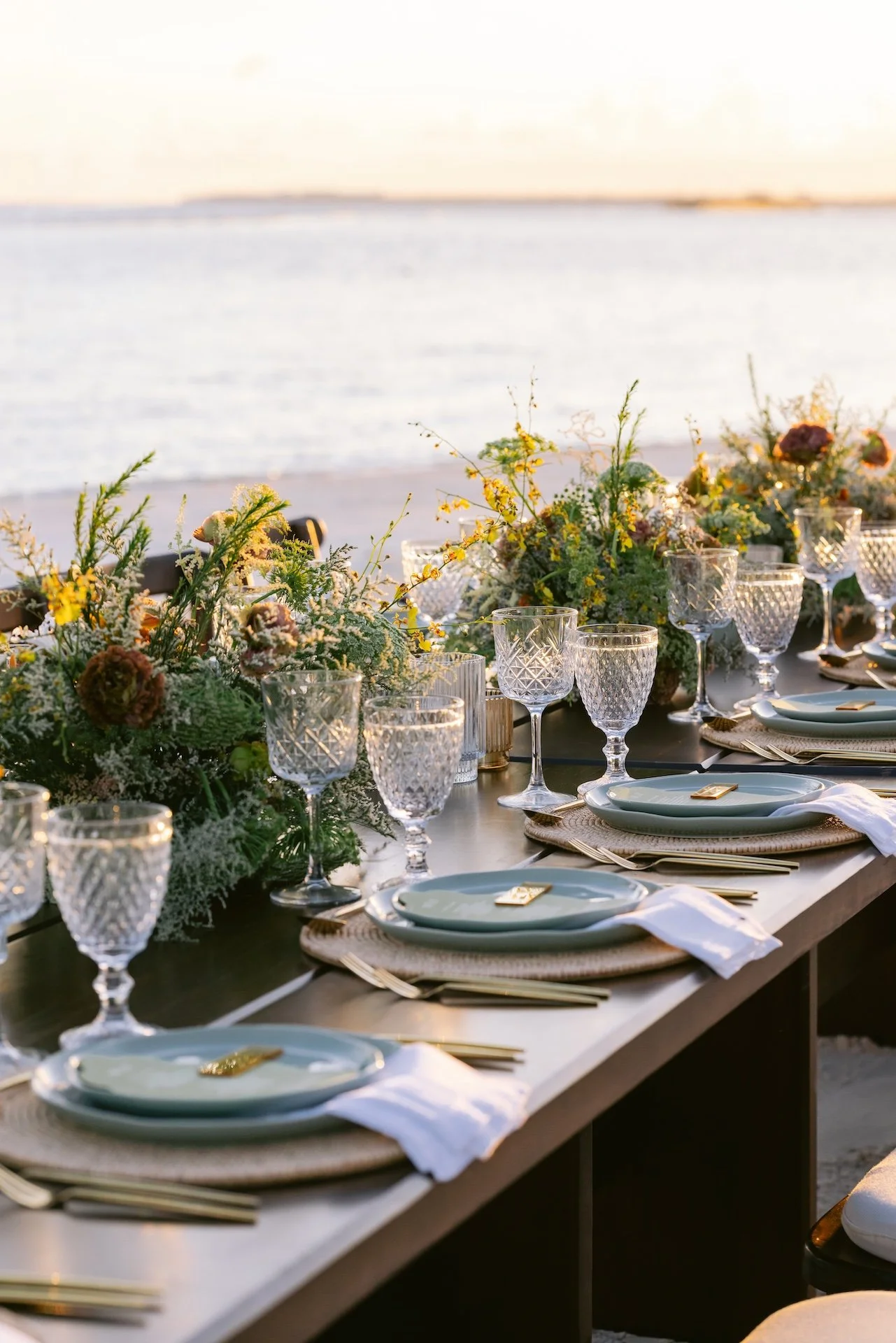A beautifully set outdoor dining table with floral centerpieces and glassware near the beach at sunset.