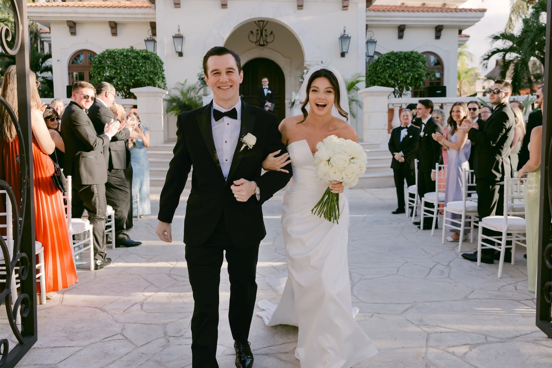 A newlywed bride and groom walk down the aisle arm in arm, smiling, as their wedding guests applaud in the background outside a white mansion with stairs and greenery.