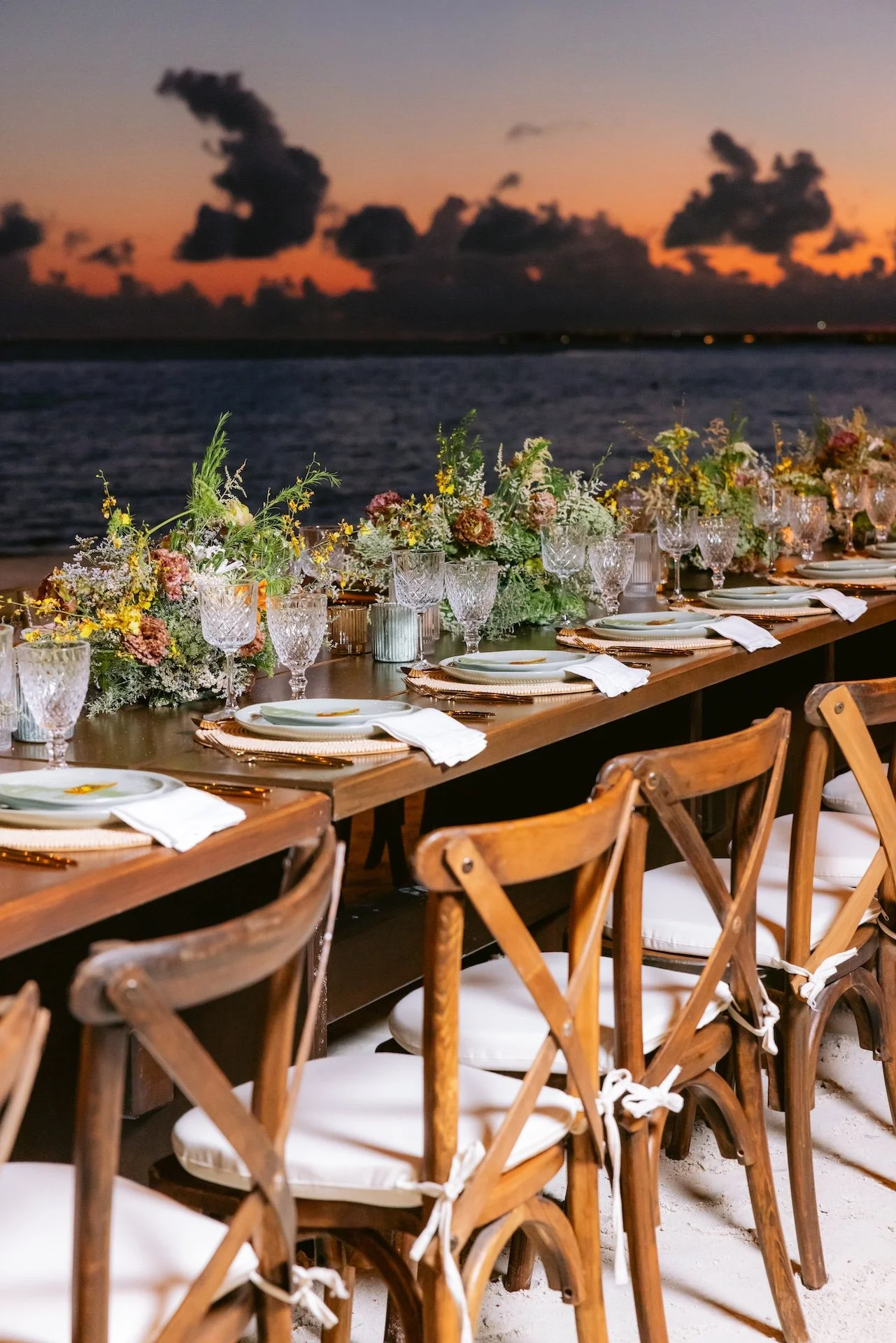Elegantly set outdoor dining table with floral arrangements, glassware, and white napkins overlooking the ocean at sunset.