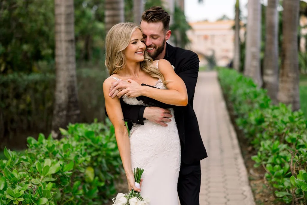 A happy couple in wedding attire embracing on a lush, green pathway lined with palm trees.