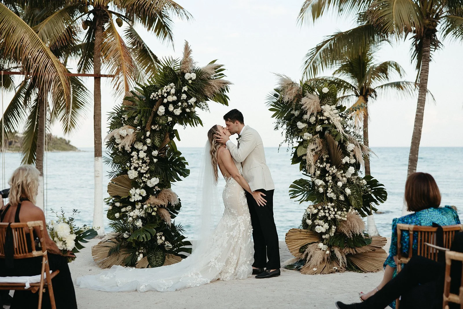 A couple kissing during a beach wedding ceremony under a floral arch with palm trees and ocean in the background.