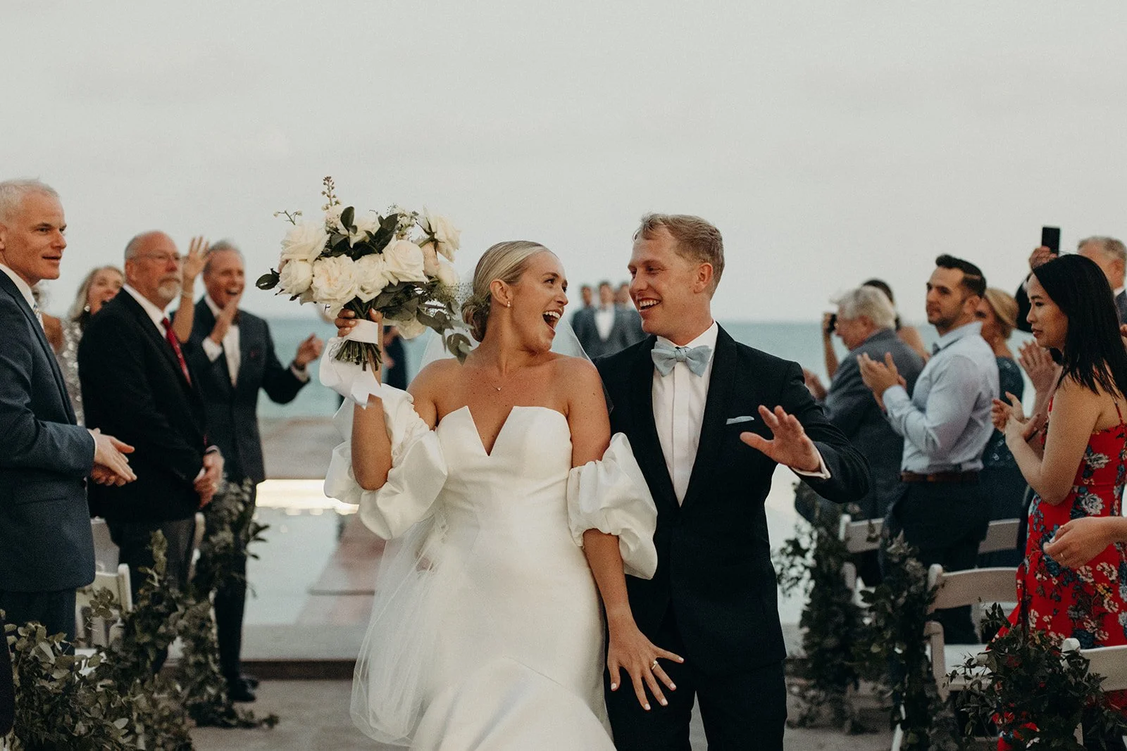 A newlywed couple walking down the aisle outdoors by the sea, smiling and celebrating with friends and family.