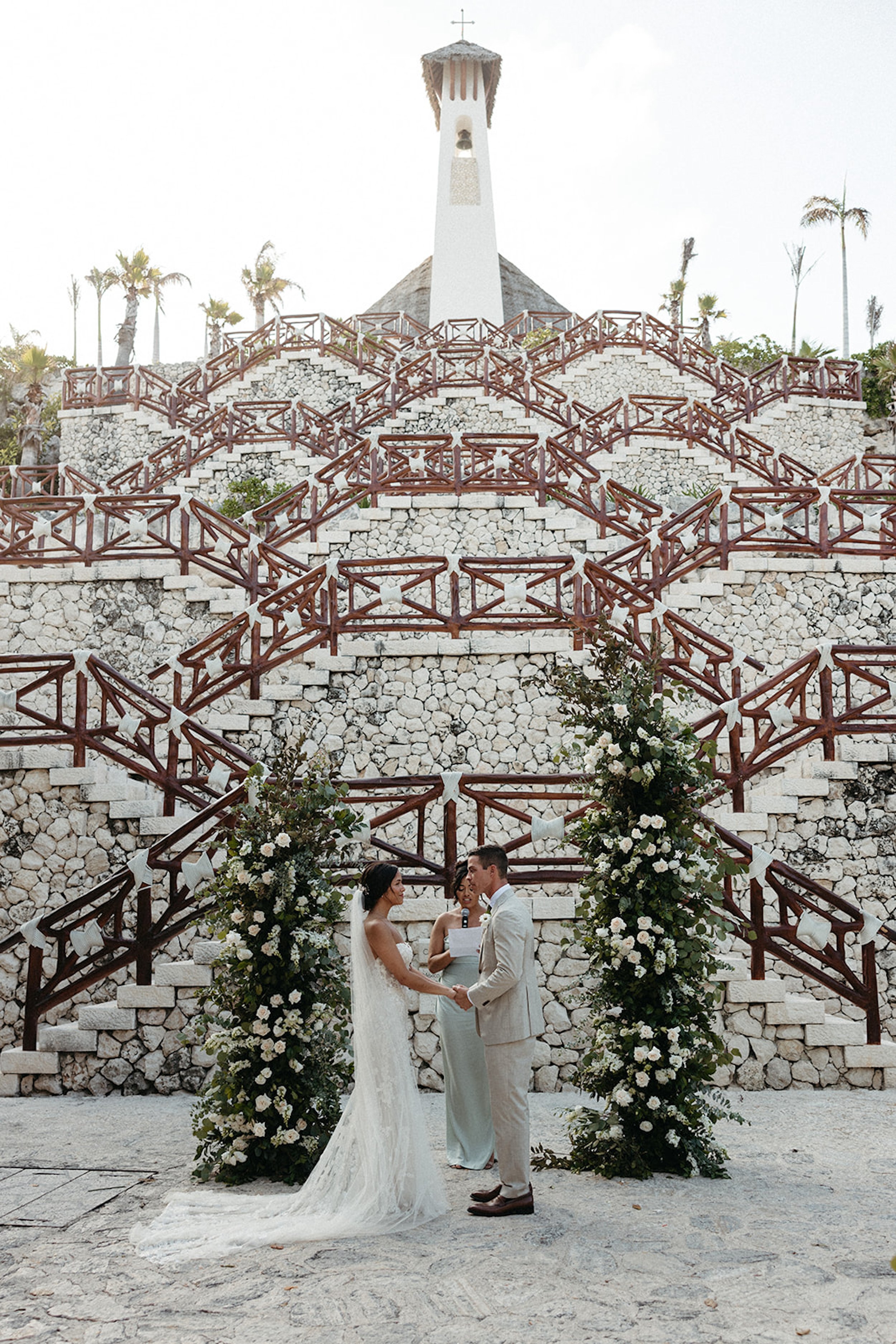 A wedding ceremony taking place outdoors in front of a stone staircase with red railings and two large floral arrangements, with a small church or chapel building with a bell tower and a cross on top in the background.