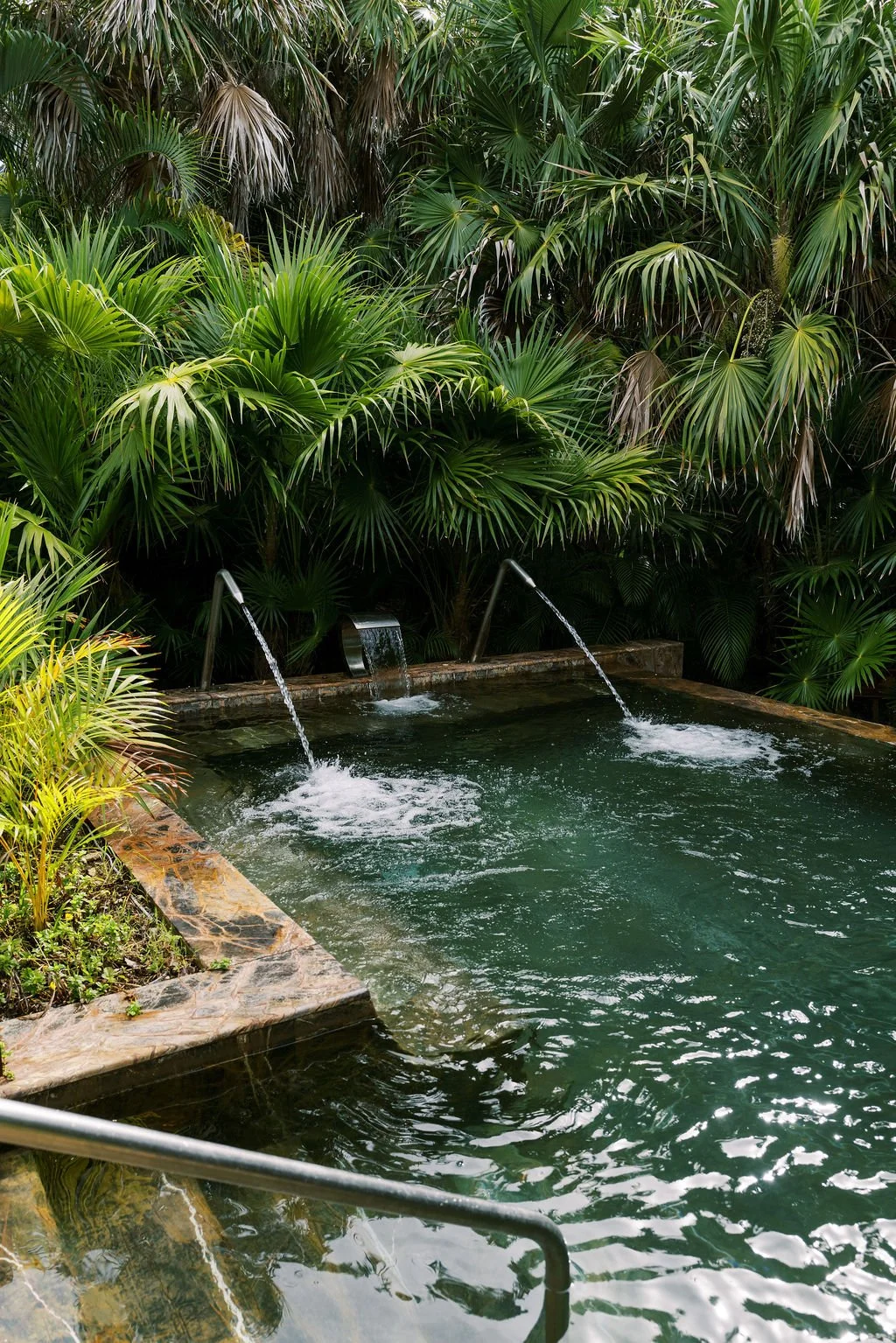 Tropical outdoor hot spring pool with water spouts, surrounded by lush green palm trees and tropical plants.
