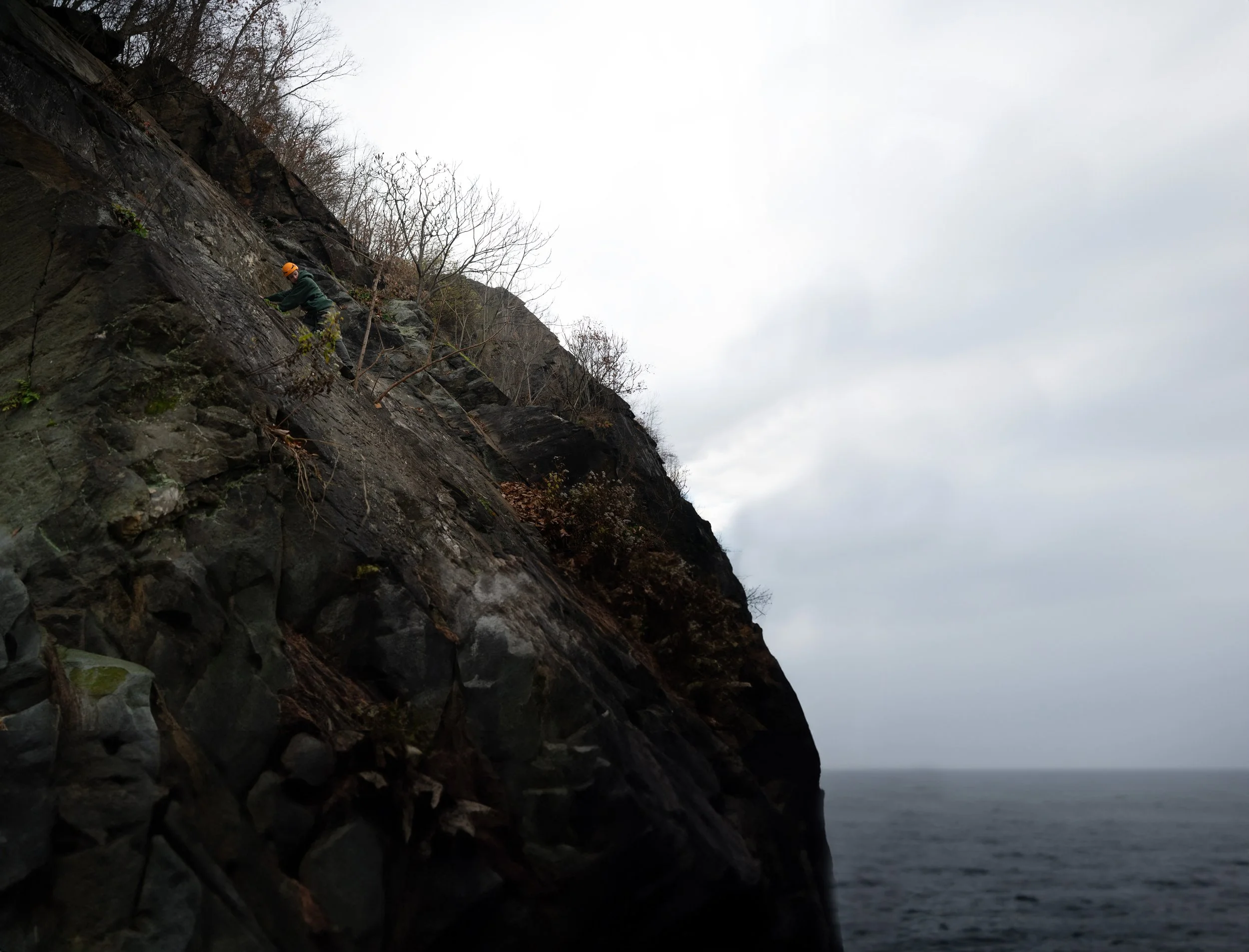 Person rock climbing on a steep cliff overlooking the ocean under a cloudy sky.
