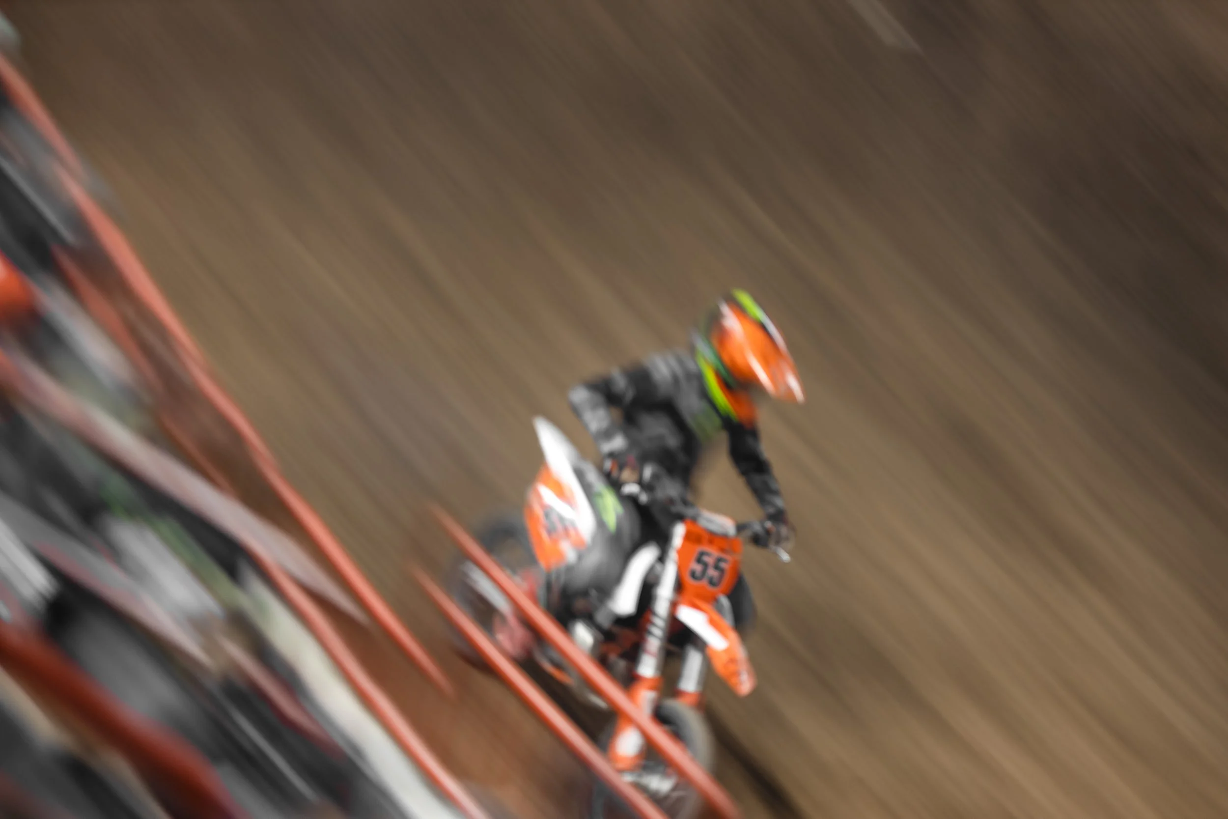 Motocross rider wearing orange helmet and riding an orange bike with the number 55, on a dirt track, captured with motion blur.