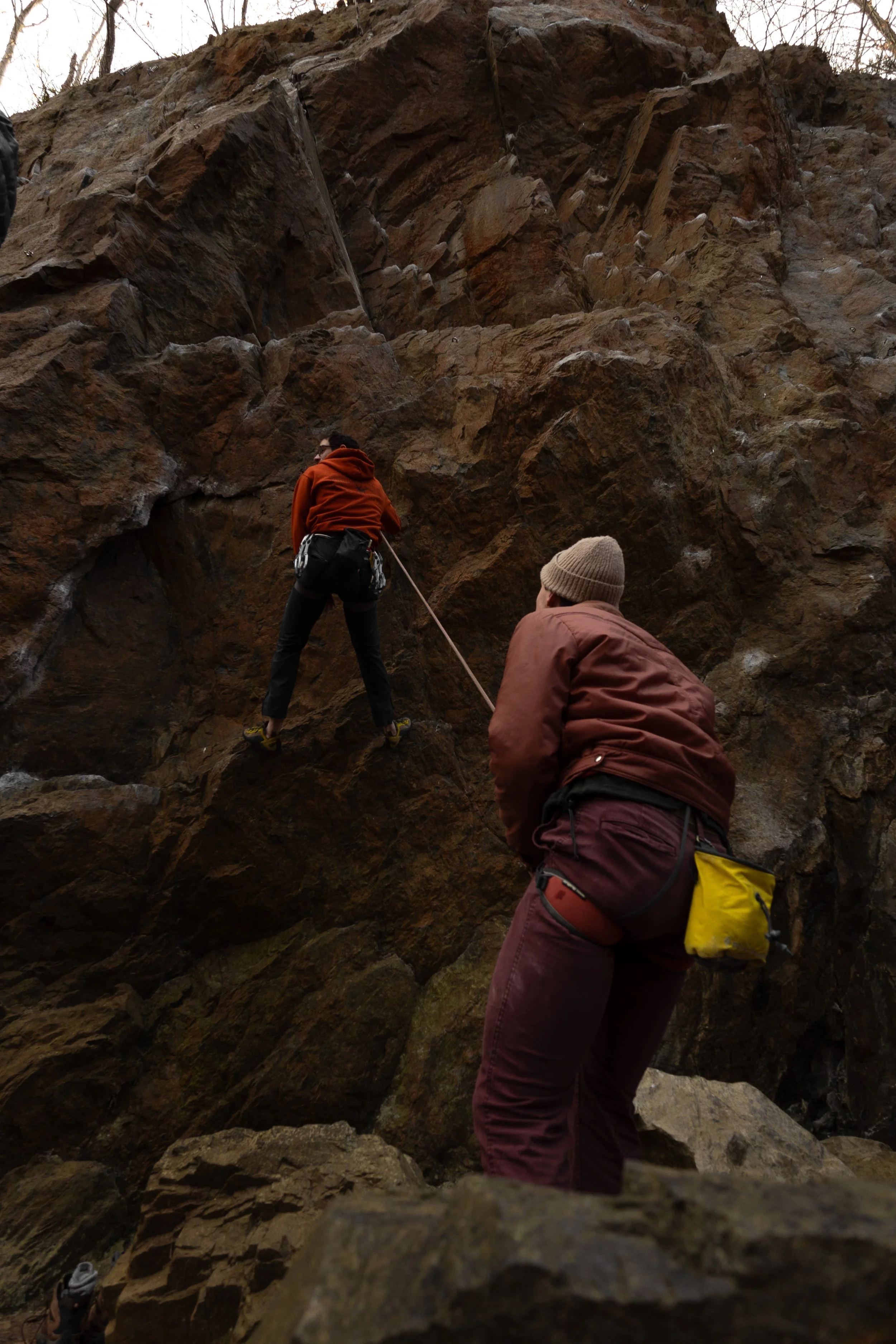 Two people practicing rock climbing outdoors on a large rocky cliff. One person is climbing the rock face, while the other is belaying from below.