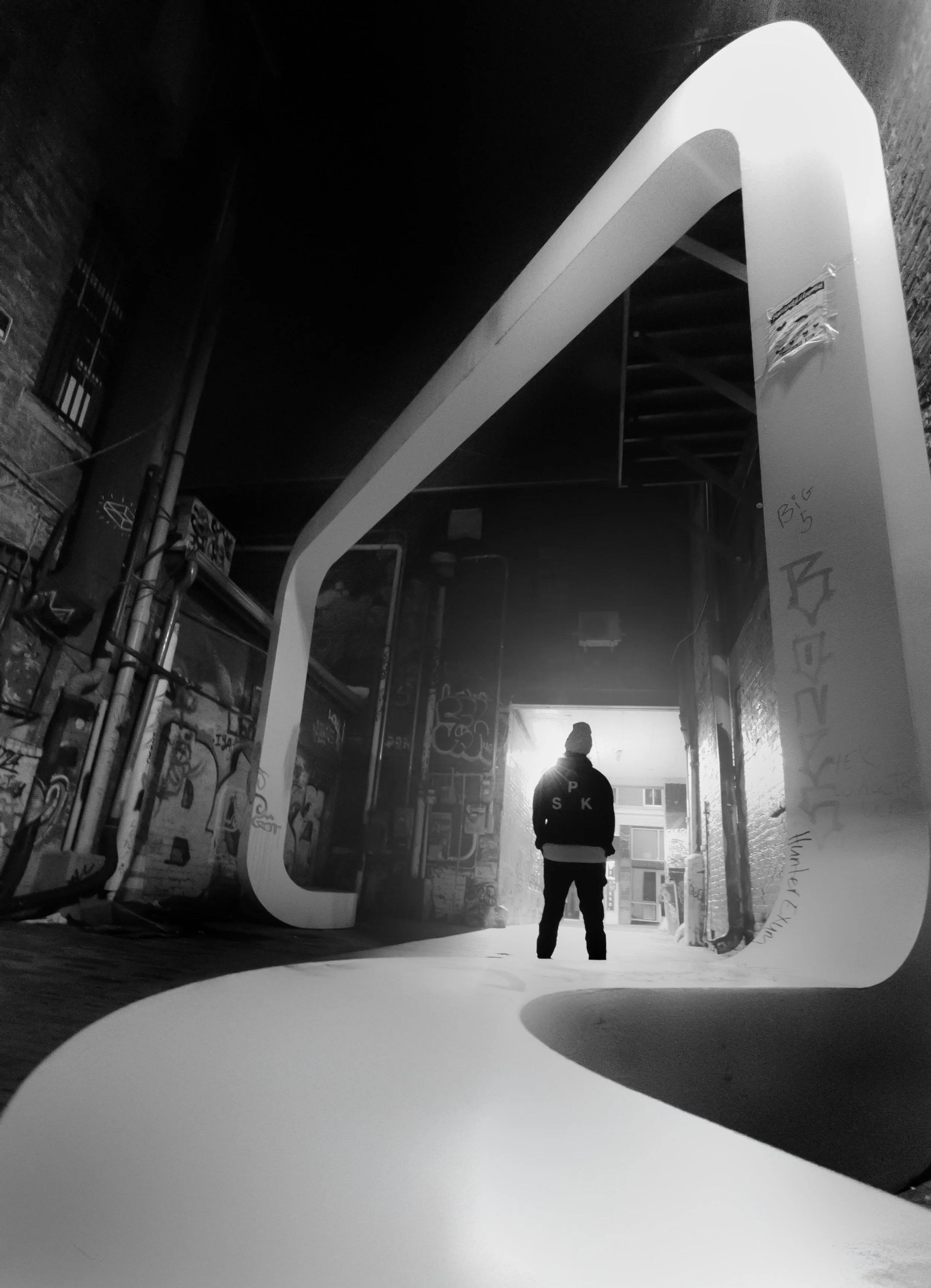 A person standing in a graffiti-covered alleyway with a large, modern, white, rectangular sculpture frame in the foreground. The person is wearing a beanie and a jacket, facing away towards a light-filled end of the alley.