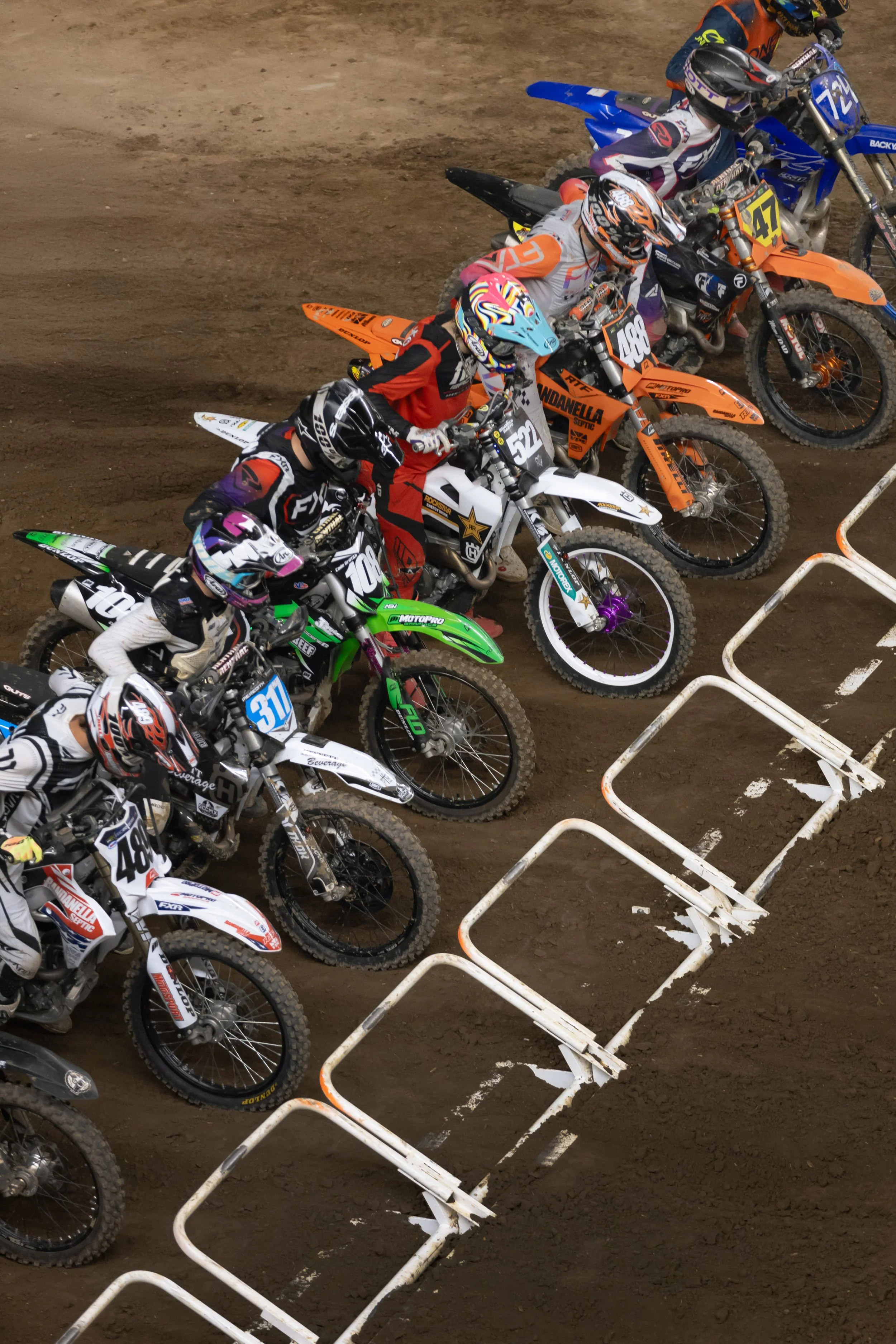 Motorcycle racers lined up at the starting line on a dirt track, wearing colorful helmets and racing gear, with a white safety barrier on the ground.