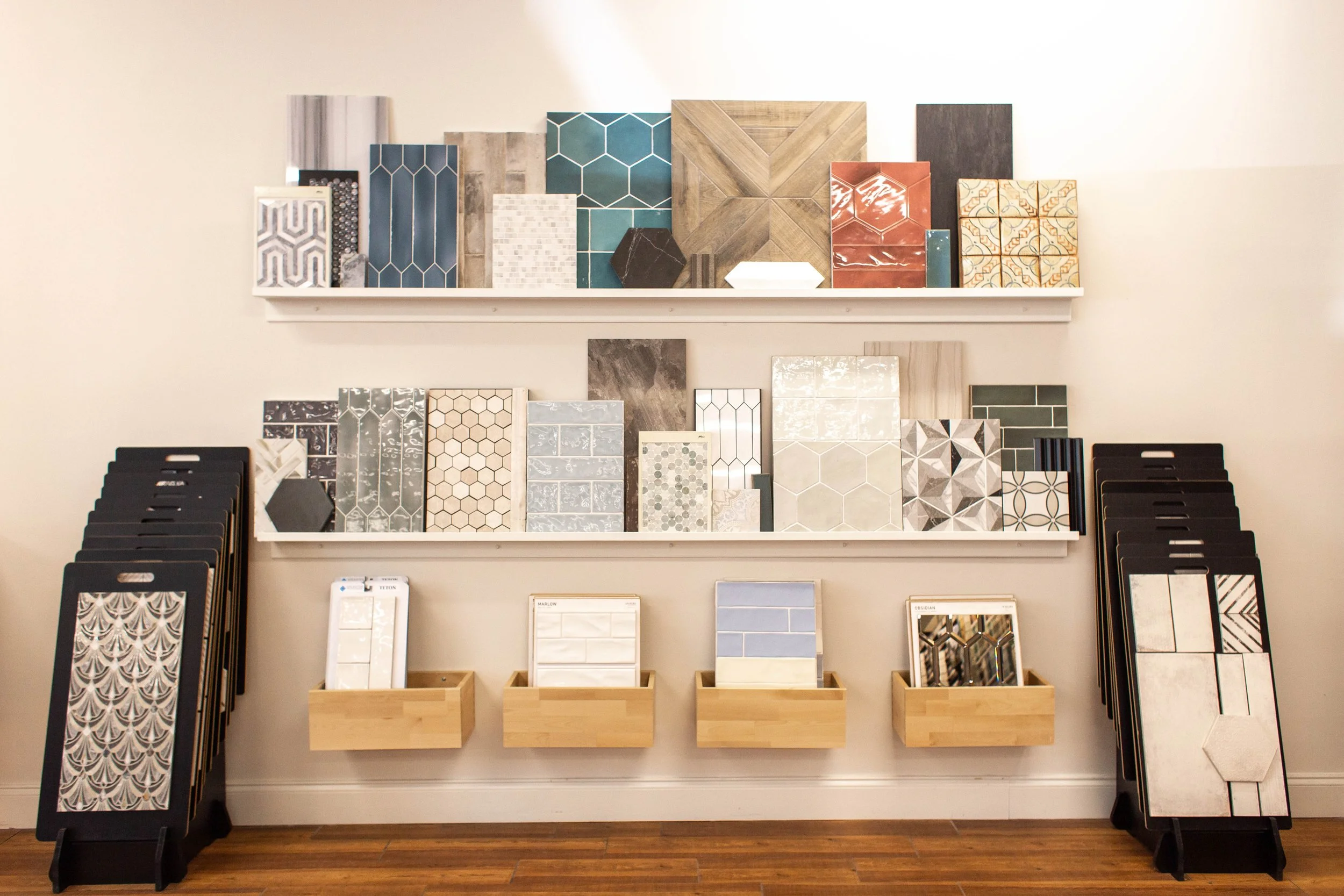 Various tile samples displayed on shelves and stands, including hexagonal, subway, and patterned designs, in a showroom setting with wooden flooring.