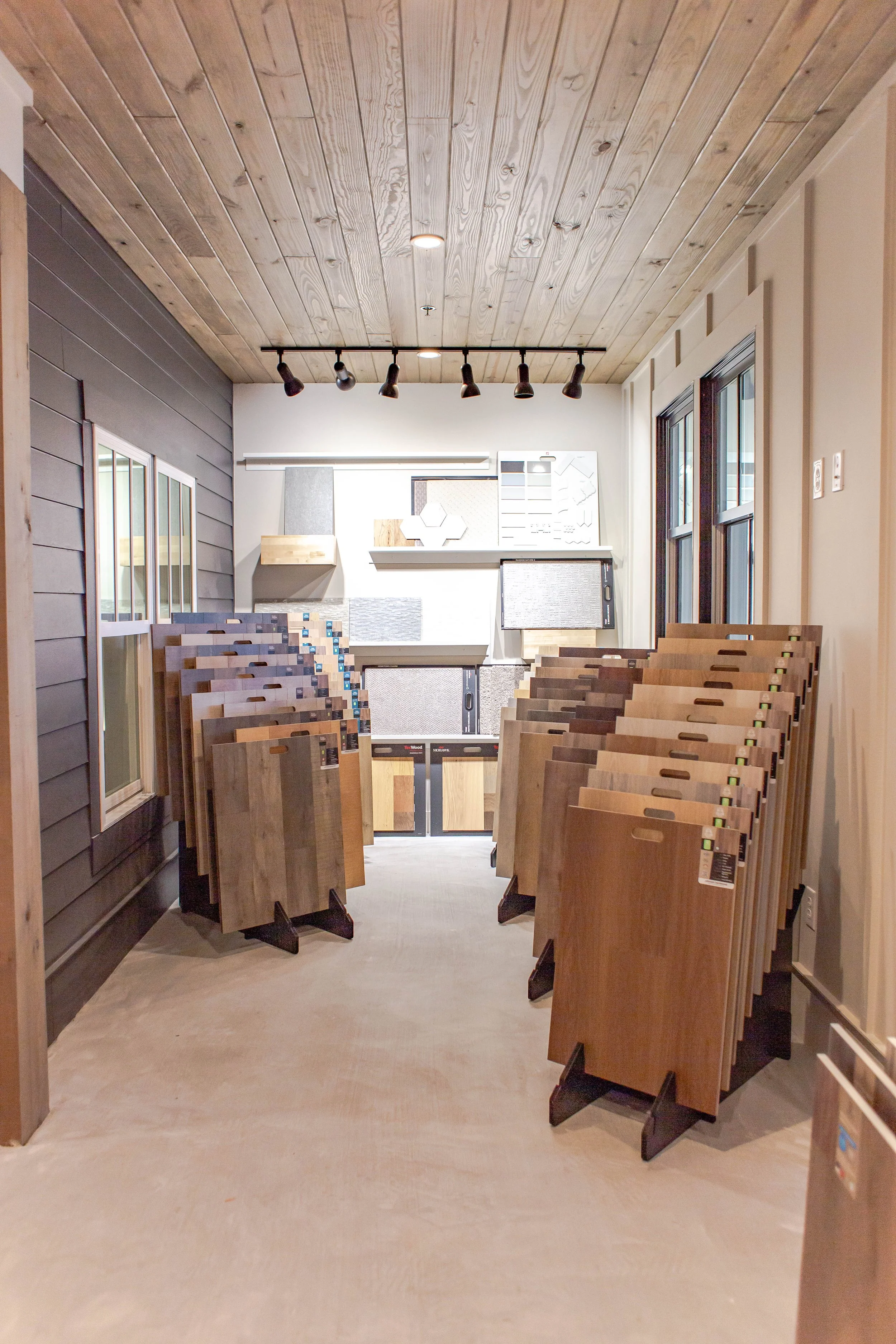 Showroom displaying various flooring samples on panels, including hardwood and laminate options. The room has a wooden ceiling and track lighting.