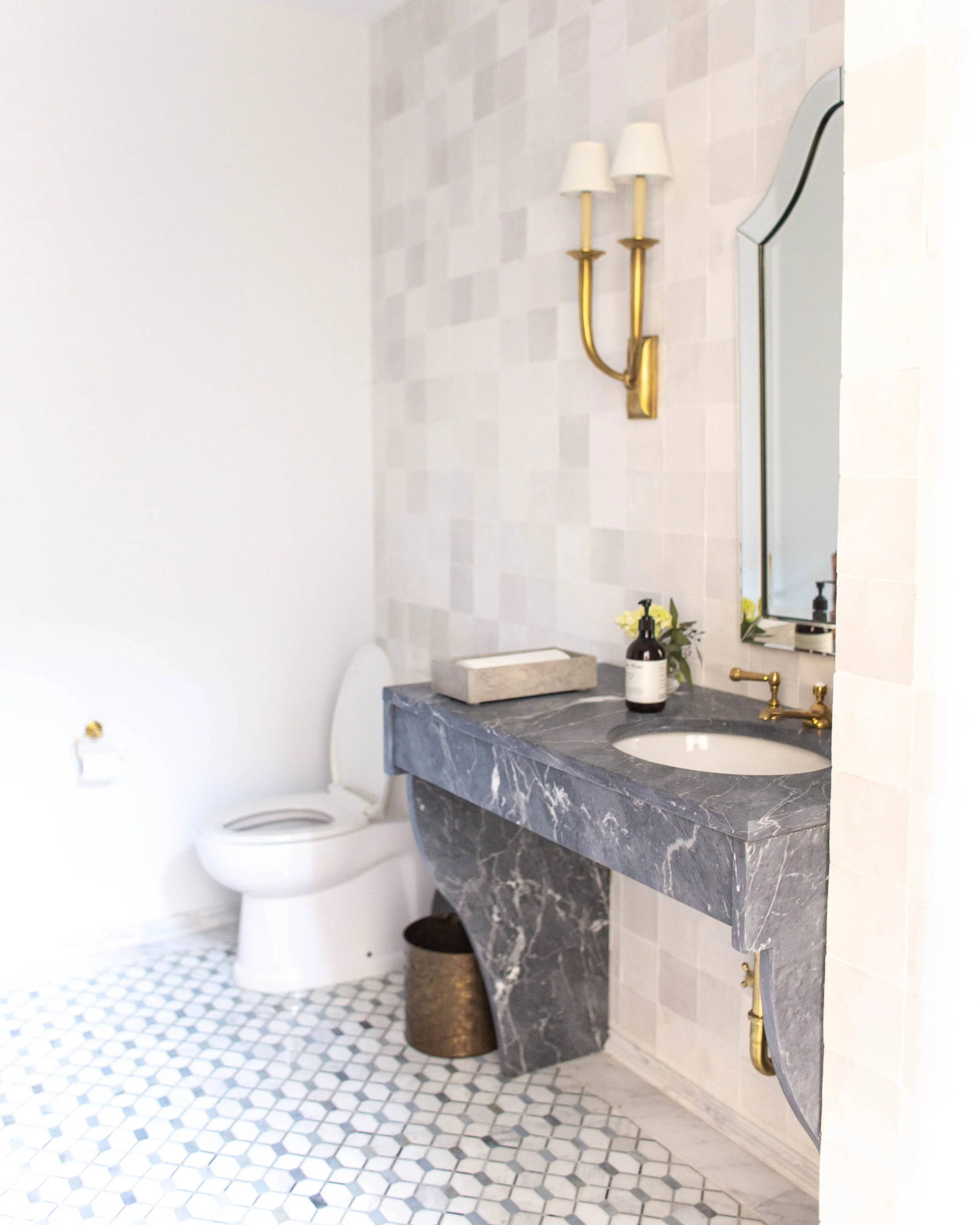 Bathroom interior with white toilet, marble countertop, gold fixtures, wall-mounted mirror, and tiled floor.