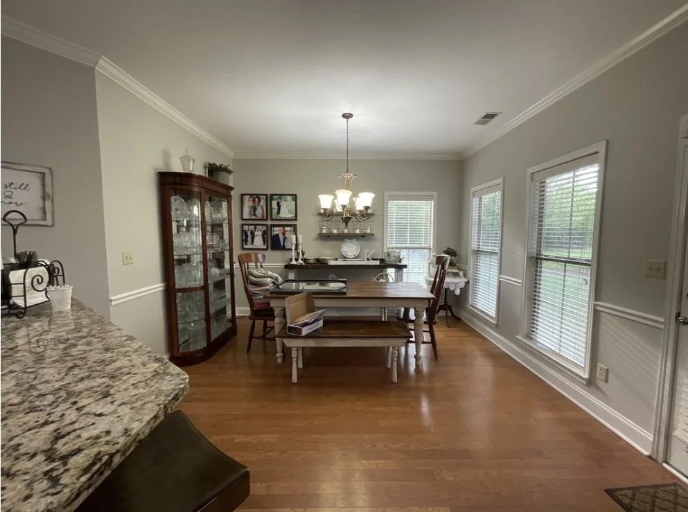 Dining room with wooden floor, table, chairs, chandelier, and large windows; glass cabinet and decorative items on walls.