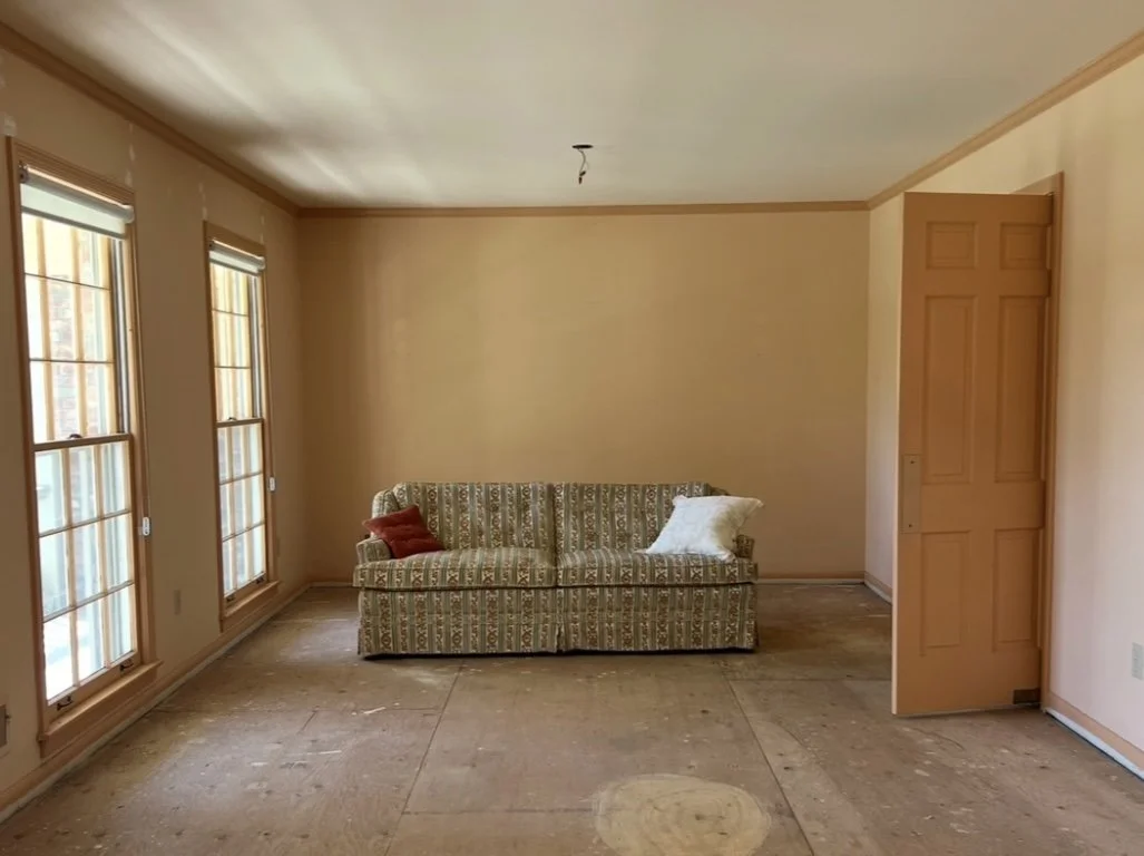 Empty room with floral-patterned sofa between two windows, beige walls, open door, and unfinished floor.