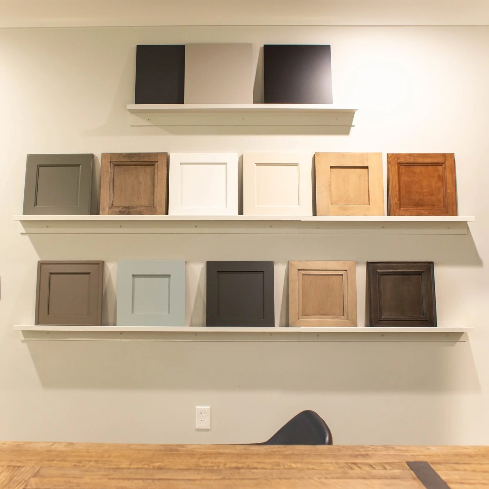 Display of various cabinet door samples mounted on a wall shelf, showcasing different designs, materials, and colors, with a wooden table in the foreground.